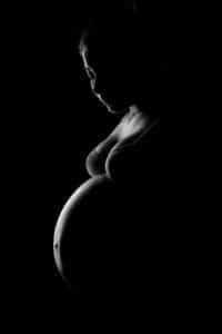 Schwarz weiss Babybauchfoto kunstvoll aufgenommen vor dunklem Hintergrund.